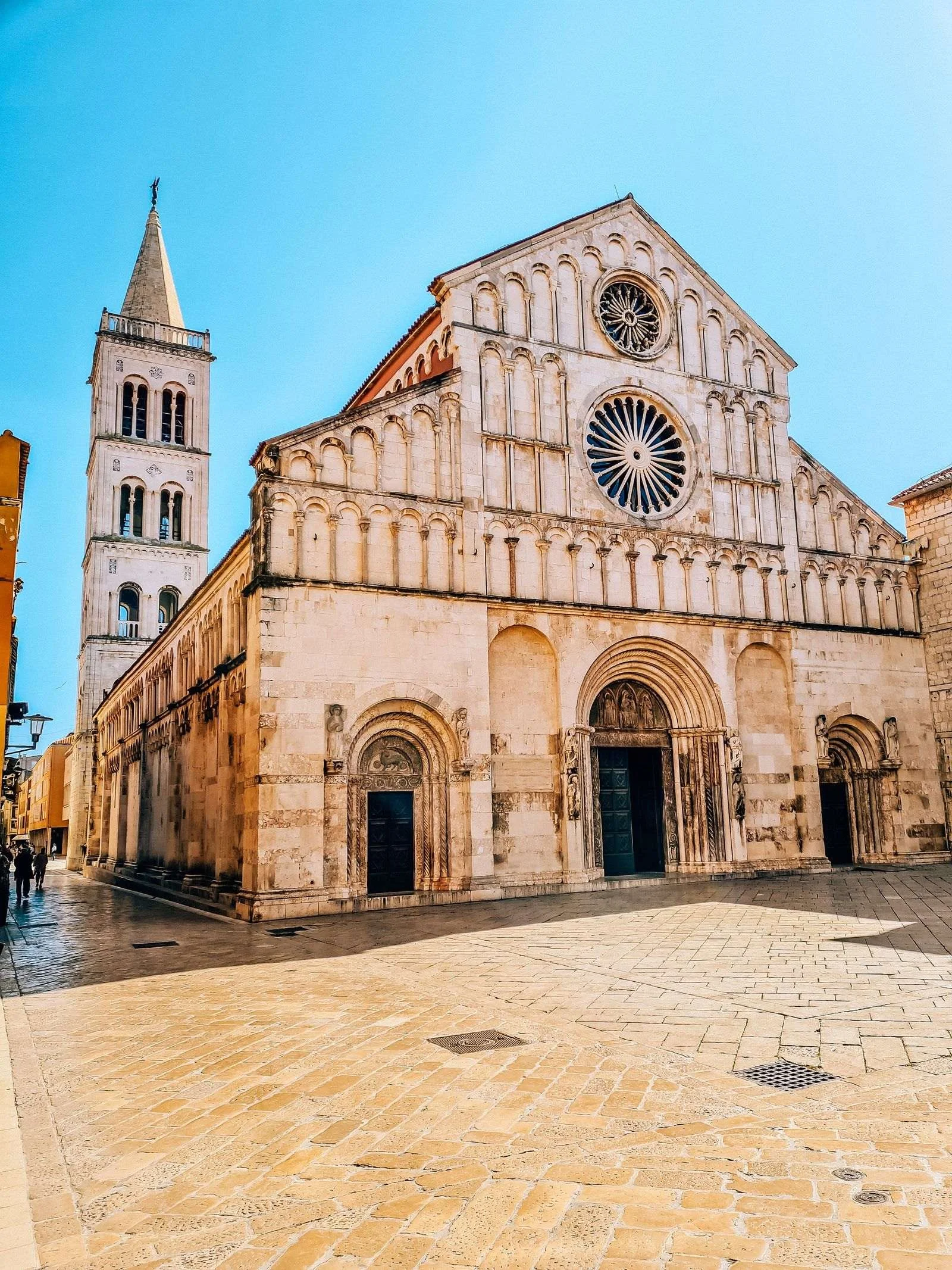 A large, light coloured stone church with carving detail on the front, 3 door ways and a large circular window at the top in the middle. a bell tower can be seen at the rear of the church in zadar