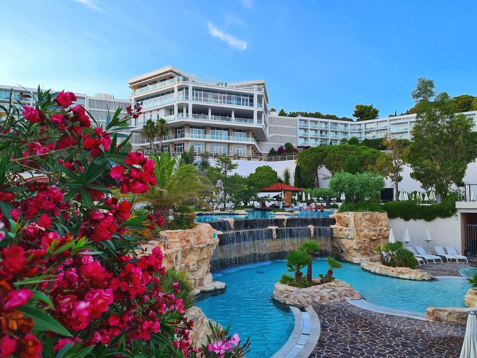 An outdoor blue pool area of an hotel. The water is connected on different levels by a waterfall. A large white hotel building is in the background with many glass windows