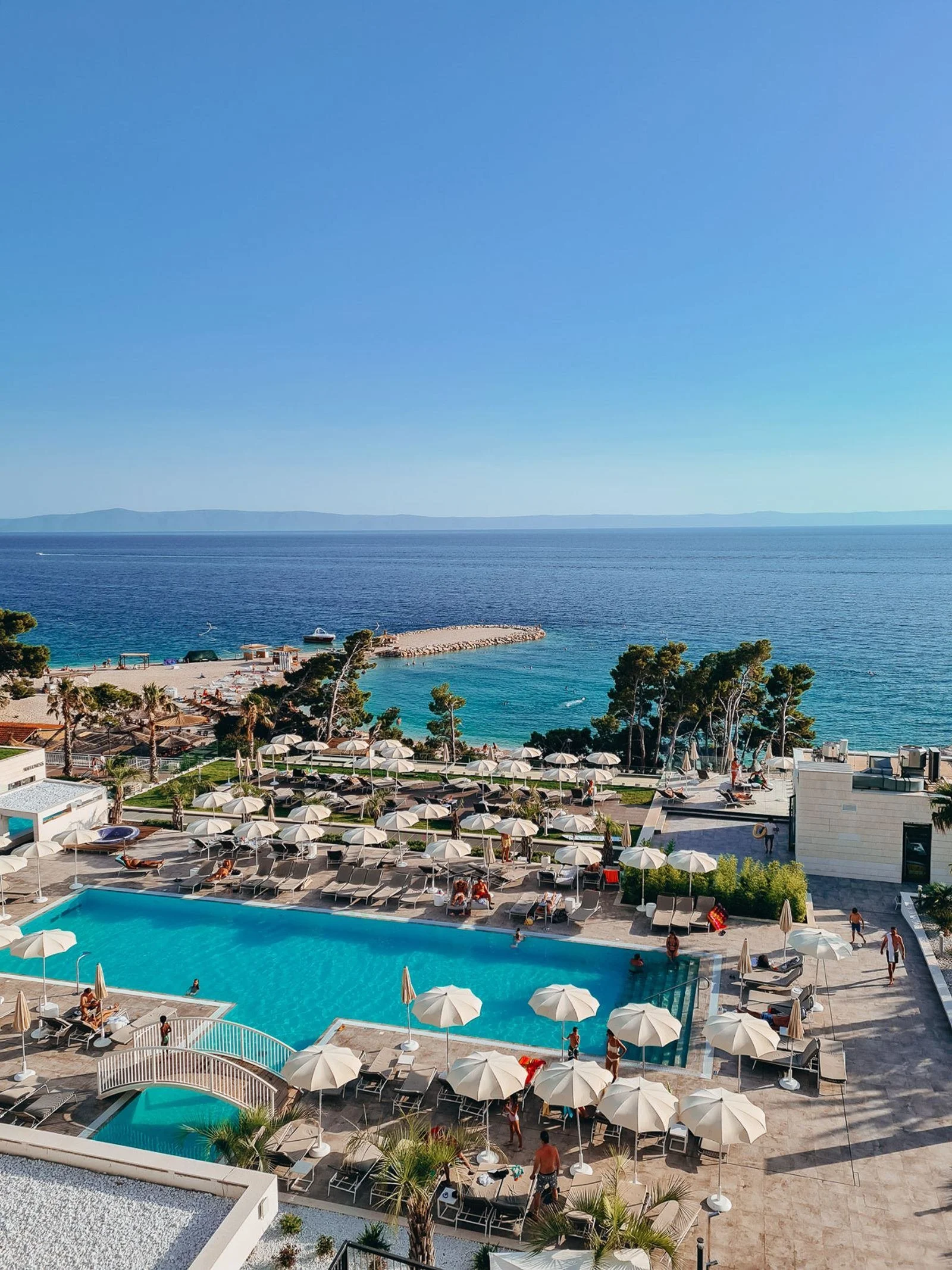 Looking down from a hotel balcony to the outdoor pool which is blue and surrounded by cream coloured parasols, the sea is beyond the pool
