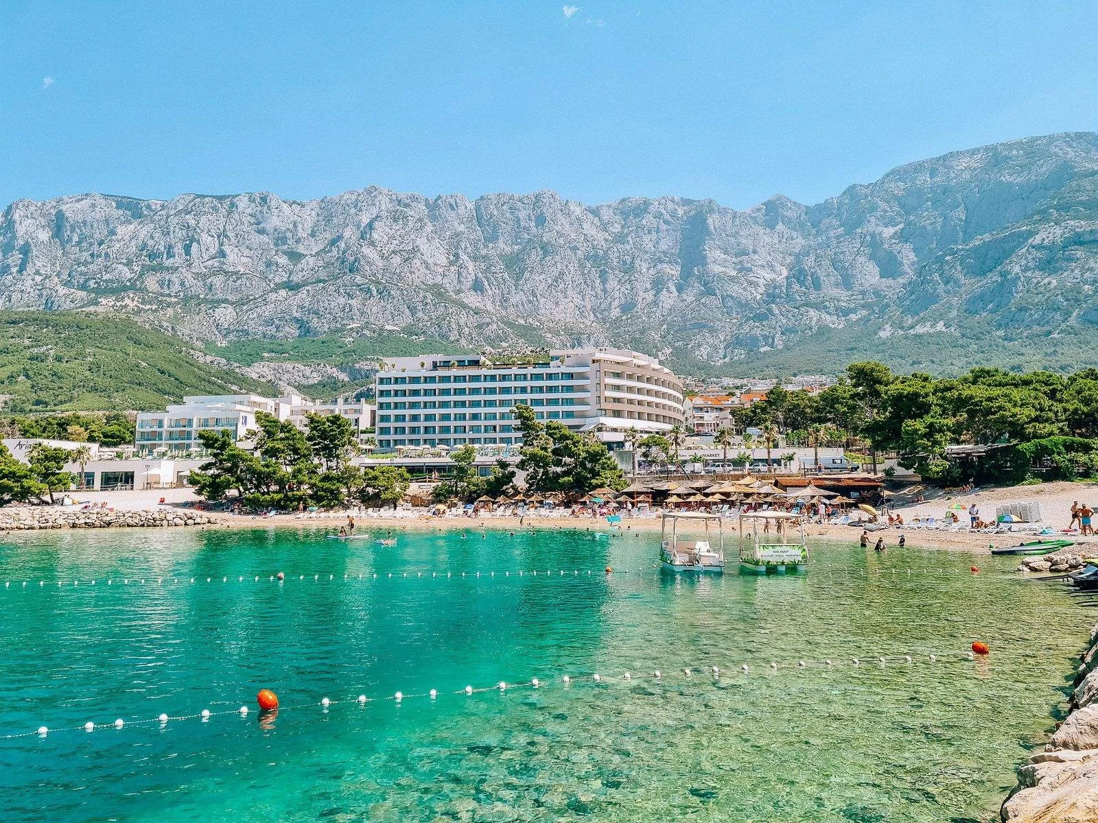 Looking across shallow, emerald green water to a beach with a 7-storymodern white hotel on it with mountains towering in the background. The beach and water is busy with some people
