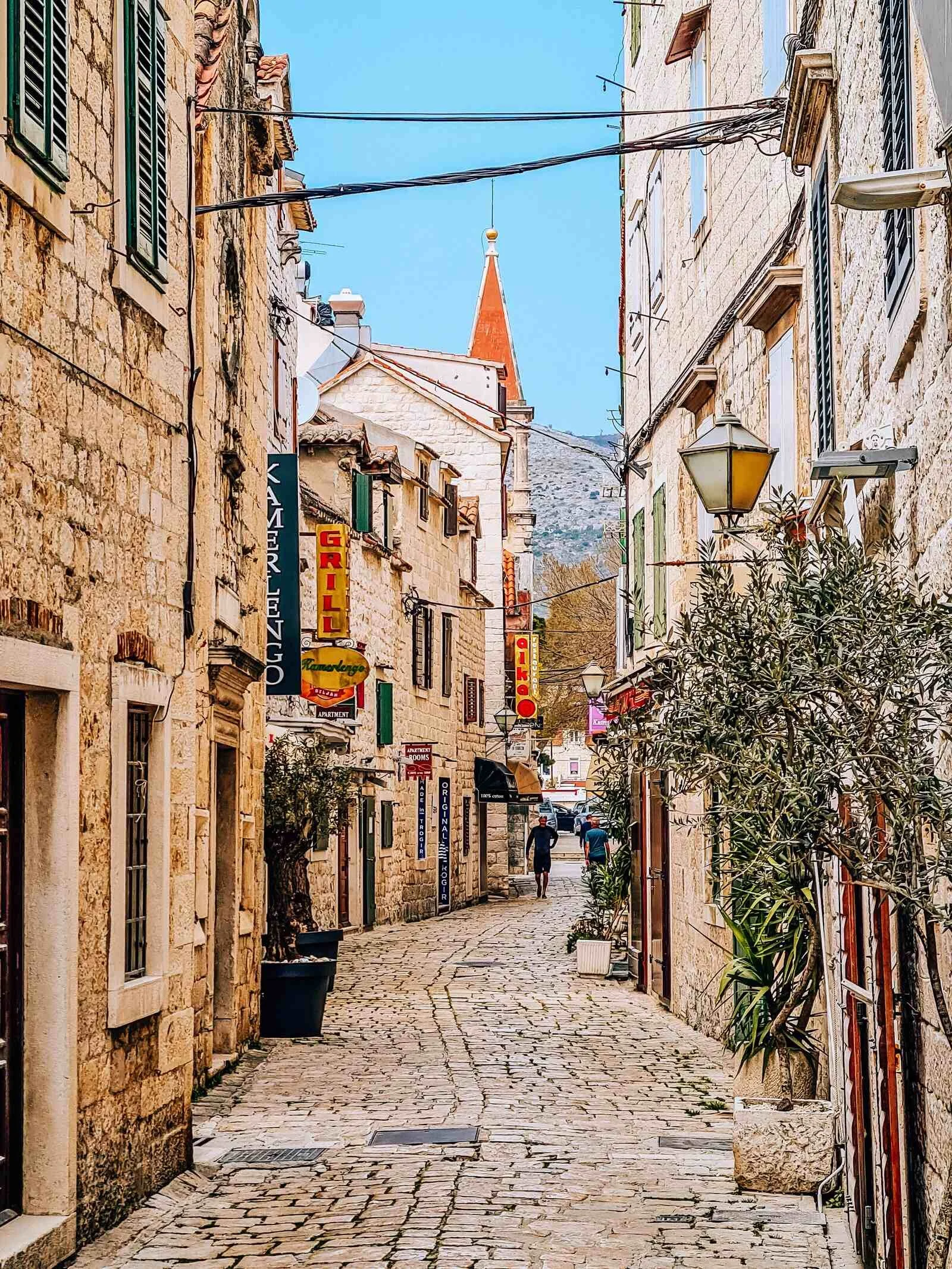 A cobbled alleyway with light stone buildings on eitherside. Shop signs and lanterns hang along the alley and a church with an orange roof spire is at the end of the alleyway. Windows with shutters overlook the alleyway all the way down